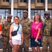 A group of seven people pose together outdoors in front of a large modern building with columns and balconies. Three individuals are wearing U.S. Army camouflage uniforms with tactical gear, while four civilians in casual summer clothing stand among them. The group appears relaxed and smiling, suggesting a friendly, informal interaction.