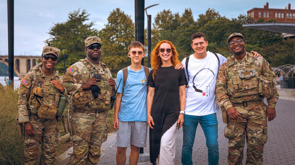 Six people pose together outdoors near a waterfront area, three of whom are wearing U.S. Army camouflage uniforms with tactical gear. The group appears to be civilians and soldiers taking a friendly photo together, likely at a public event or tourist location. The setting features trees, a bridge, and a brick building visible in the background.