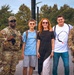 Six people pose together outdoors near a waterfront area, three of whom are wearing U.S. Army camouflage uniforms with tactical gear. The group appears to be civilians and soldiers taking a friendly photo together, likely at a public event or tourist location. The setting features trees, a bridge, and a brick building visible in the background.