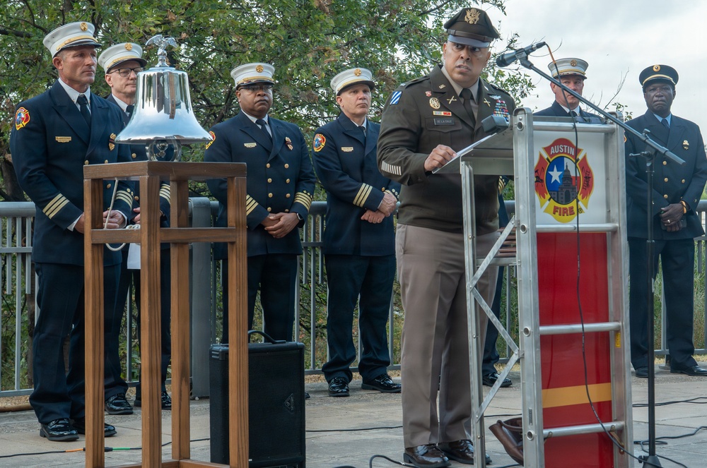 A formal outdoor ceremony featuring an Army officer in olive/brown dress uniform speaking at a podium bearing the Austin Fire Department insignia. Several fire department officials in dark dress uniforms with white caps stand in the background near a ceremonial silver bell mounted on a wooden stand. The event appears to be a memorial or commemorative ceremony, likely related to first responders.