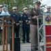 A formal outdoor ceremony featuring an Army officer in olive/brown dress uniform speaking at a podium bearing the Austin Fire Department insignia. Several fire department officials in dark dress uniforms with white caps stand in the background near a ceremonial silver bell mounted on a wooden stand. The event appears to be a memorial or commemorative ceremony, likely related to first responders.