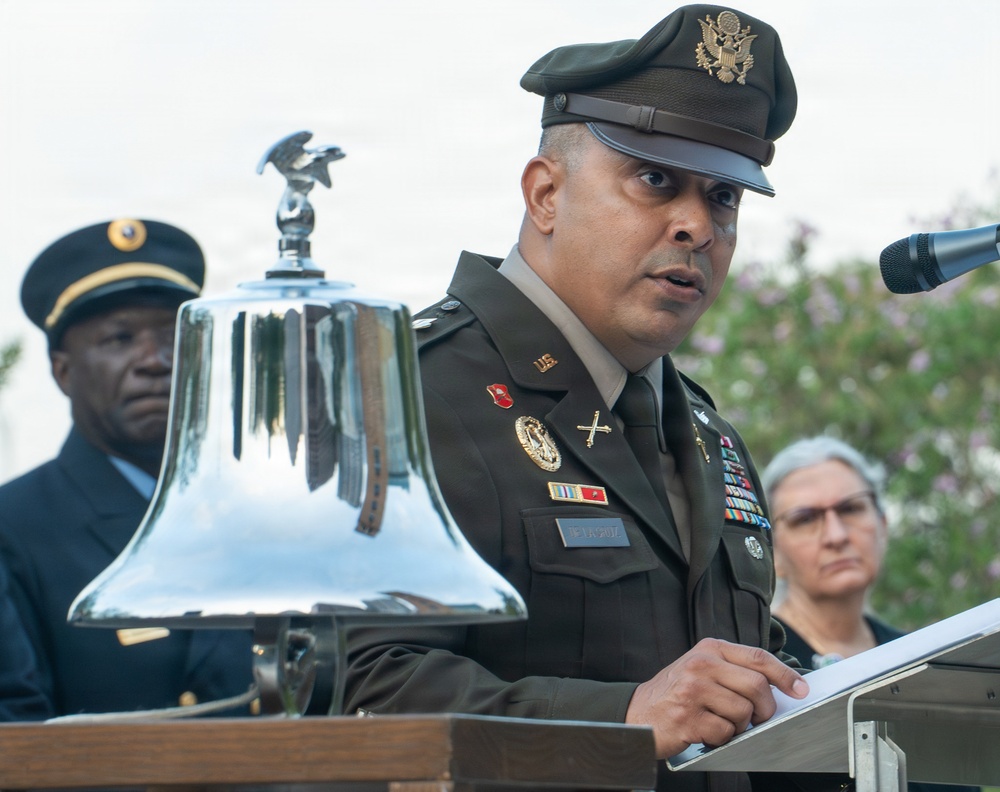 A U.S. Army officer in dress green uniform is speaking at a podium during what appears to be a formal military ceremony. A polished ceremonial bell with an eagle finial is prominently displayed on the podium. Two other individuals are visible in the background, one in military uniform and one in civilian attire.
