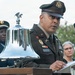 A U.S. Army officer in dress green uniform is speaking at a podium during what appears to be a formal military ceremony. A polished ceremonial bell with an eagle finial is prominently displayed on the podium. Two other individuals are visible in the background, one in military uniform and one in civilian attire.