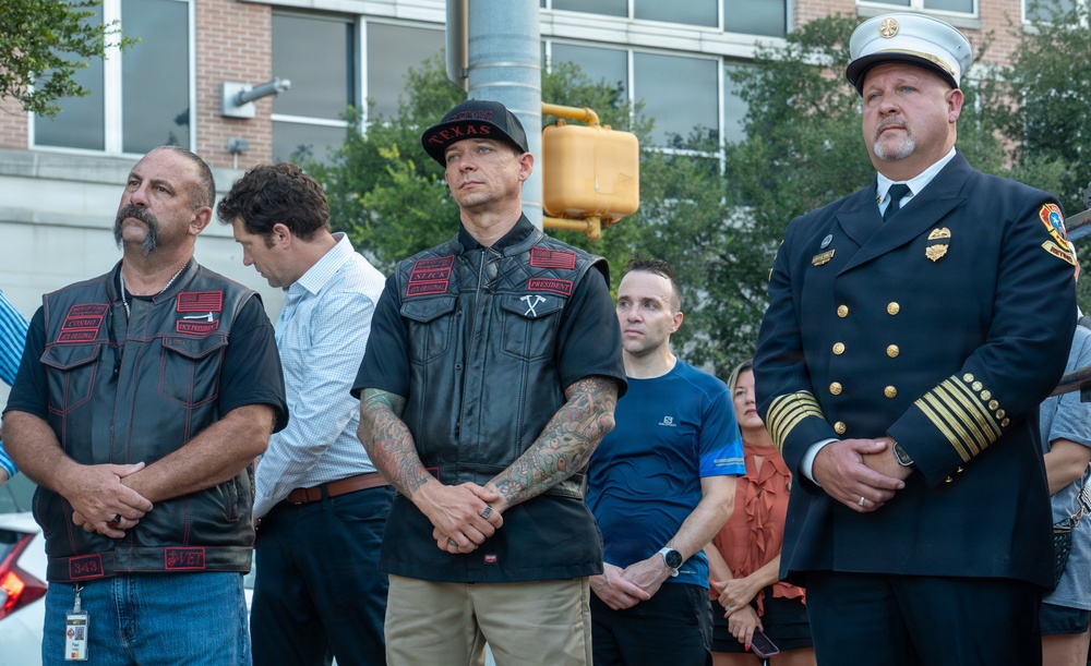 A group of individuals stand together at what appears to be an outdoor memorial or ceremonial event. The group includes two men wearing black leather vests with patches, one wearing a 'Texas' cap, and a fire department official in full dress uniform with gold insignia. The solemn postures and clasped hands suggest a moment of remembrance or tribute.