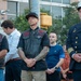 A group of individuals stand together at what appears to be an outdoor memorial or ceremonial event. The group includes two men wearing black leather vests with patches, one wearing a 'Texas' cap, and a fire department official in full dress uniform with gold insignia. The solemn postures and clasped hands suggest a moment of remembrance or tribute.