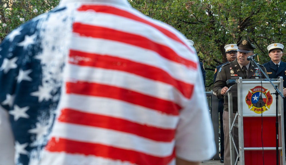 A military officer in Army dress uniform speaks at a podium bearing the Austin Fire Department insignia, while an attendee wearing an American flag-patterned shirt stands in the foreground with their back to the camera. Additional uniformed personnel, including what appears to be a fire department officer in white dress uniform, stand nearby. The scene appears to be a formal public ceremony, likely a memorial or civic event.