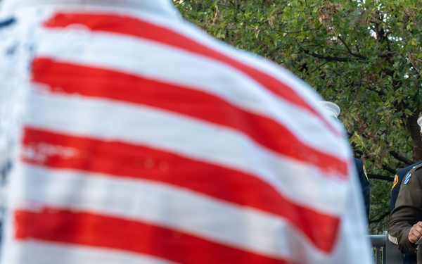 A military officer in Army dress uniform speaks at a podium bearing the Austin Fire Department insignia, while an attendee wearing an American flag-patterned shirt stands in the foreground with their back to the camera. Additional uniformed personnel, including what appears to be a fire department officer in white dress uniform, stand nearby. The scene appears to be a formal public ceremony, likely a memorial or civic event.