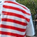 A military officer in Army dress uniform speaks at a podium bearing the Austin Fire Department insignia, while an attendee wearing an American flag-patterned shirt stands in the foreground with their back to the camera. Additional uniformed personnel, including what appears to be a fire department officer in white dress uniform, stand nearby. The scene appears to be a formal public ceremony, likely a memorial or civic event.
