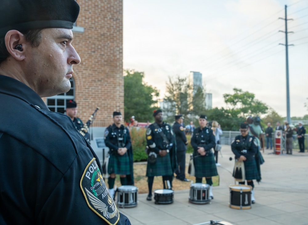 A police pipe and drum band is assembled outdoors, likely for a ceremonial event. Officers wearing dark uniforms with green tartan kilts stand at attention with snare drums positioned on the ground before them. The foreground features a close-up profile of a uniformed officer wearing an Austin Police Department patch and a black beret with an in-ear monitor.