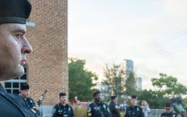 A police pipe and drum band is assembled outdoors, likely for a ceremonial event. Officers wearing dark uniforms with green tartan kilts stand at attention with snare drums positioned on the ground before them. The foreground features a close-up profile of a uniformed officer wearing an Austin Police Department patch and a black beret with an in-ear monitor.