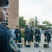 A police pipe and drum band is assembled outdoors, likely for a ceremonial event. Officers wearing dark uniforms with green tartan kilts stand at attention with snare drums positioned on the ground before them. The foreground features a close-up profile of a uniformed officer wearing an Austin Police Department patch and a black beret with an in-ear monitor.