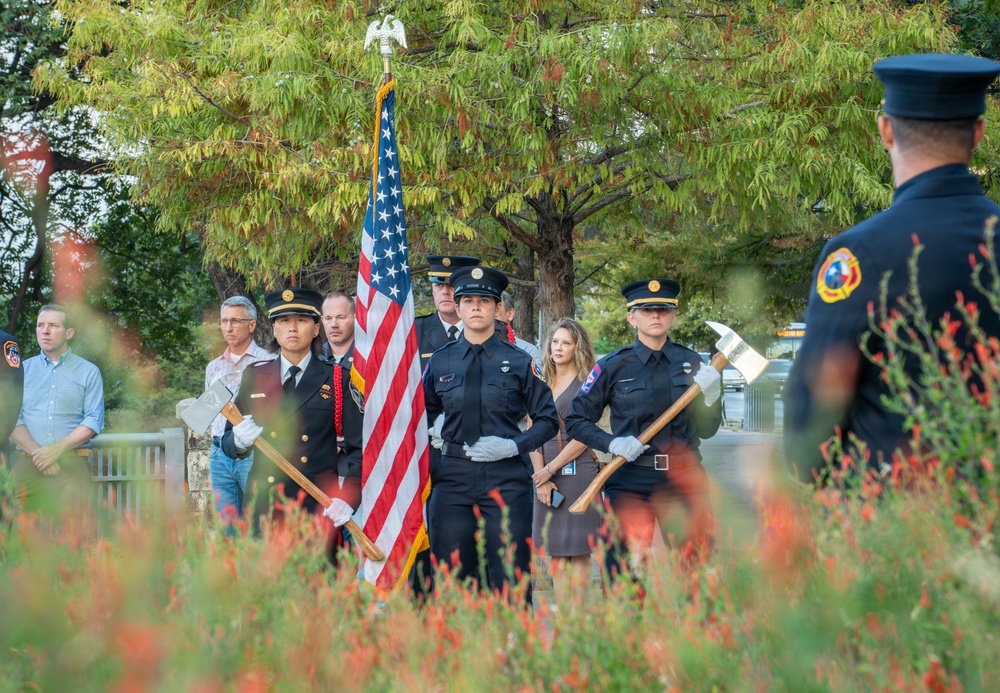 A firefighter color guard ceremony is taking place outdoors, with uniformed firefighters carrying an American flag and ceremonial axes. The formation appears to be part of a formal memorial or commemorative event, likely a 9/11 remembrance given the firefighter uniforms and ceremonial nature. The scene is photographed through blooming red wildflowers in the foreground, with civilian spectators visible in the background.
