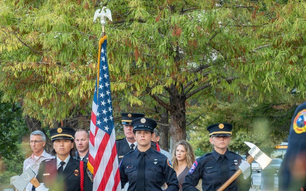 A firefighter color guard ceremony is taking place outdoors, with uniformed firefighters carrying an American flag and ceremonial axes. The formation appears to be part of a formal memorial or commemorative event, likely a 9/11 remembrance given the firefighter uniforms and ceremonial nature. The scene is photographed through blooming red wildflowers in the foreground, with civilian spectators visible in the background.