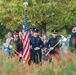 A firefighter color guard ceremony is taking place outdoors, with uniformed firefighters carrying an American flag and ceremonial axes. The formation appears to be part of a formal memorial or commemorative event, likely a 9/11 remembrance given the firefighter uniforms and ceremonial nature. The scene is photographed through blooming red wildflowers in the foreground, with civilian spectators visible in the background.