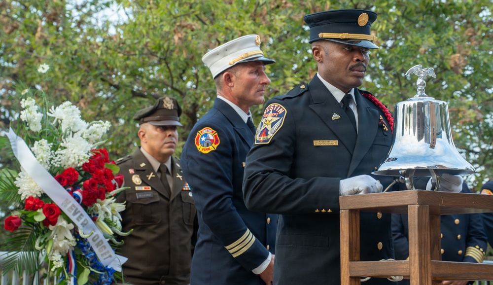 A solemn memorial ceremony featuring Austin Fire Department personnel and a U.S. Army officer. A firefighter in dress uniform is positioned at a ceremonial silver bell on a wooden stand, while a white-helmeted fire official and a uniformed soldier stand in the background. A floral wreath with red, white, and blue flowers bearing a 'Never Forget' ribbon is visible to the left.