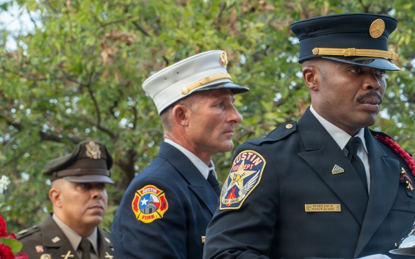 A solemn memorial ceremony featuring Austin Fire Department personnel and a U.S. Army officer. A firefighter in dress uniform is positioned at a ceremonial silver bell on a wooden stand, while a white-helmeted fire official and a uniformed soldier stand in the background. A floral wreath with red, white, and blue flowers bearing a 'Never Forget' ribbon is visible to the left.