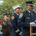 A solemn memorial ceremony featuring Austin Fire Department personnel and a U.S. Army officer. A firefighter in dress uniform is positioned at a ceremonial silver bell on a wooden stand, while a white-helmeted fire official and a uniformed soldier stand in the background. A floral wreath with red, white, and blue flowers bearing a 'Never Forget' ribbon is visible to the left.