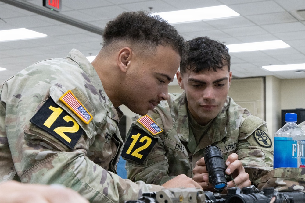 Two U.S. Army soldiers wearing MultiCam (OCP) uniforms and numbered competition bibs (both labeled '12') are closely examining a rifle scope or optical device. The soldier on the right, identified by a name tape reading 'DIAZ' and a U.S. Army tape, is holding the optic while both inspect it. The setting appears to be an indoor facility, likely during a military skills competition or training event.
