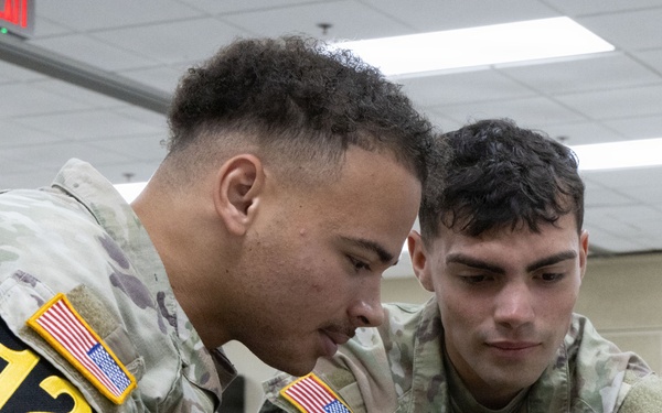 Two U.S. Army soldiers wearing MultiCam (OCP) uniforms and numbered competition bibs (both labeled '12') are closely examining a rifle scope or optical device. The soldier on the right, identified by a name tape reading 'DIAZ' and a U.S. Army tape, is holding the optic while both inspect it. The setting appears to be an indoor facility, likely during a military skills competition or training event.