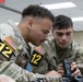 Two U.S. Army soldiers wearing MultiCam (OCP) uniforms and numbered competition bibs (both labeled '12') are closely examining a rifle scope or optical device. The soldier on the right, identified by a name tape reading 'DIAZ' and a U.S. Army tape, is holding the optic while both inspect it. The setting appears to be an indoor facility, likely during a military skills competition or training event.