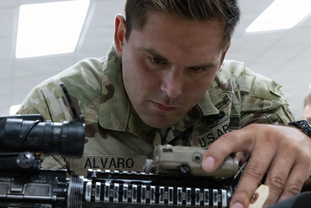 A U.S. Army soldier named Alvaro is closely inspecting or performing maintenance on a military rifle, likely an M4 or similar platform. The soldier is wearing Operational Camouflage Pattern (OCP) uniform with a 'U.S. ARMY' tape and name tape reading 'ALVARO' clearly visible. The image is a close-up shot taken in what appears to be an indoor facility with overhead lighting.