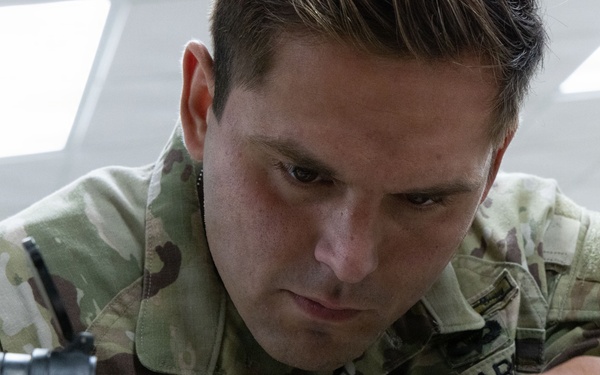 A U.S. Army soldier named Alvaro is closely inspecting or performing maintenance on a military rifle, likely an M4 or similar platform. The soldier is wearing Operational Camouflage Pattern (OCP) uniform with a 'U.S. ARMY' tape and name tape reading 'ALVARO' clearly visible. The image is a close-up shot taken in what appears to be an indoor facility with overhead lighting.