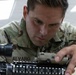 A U.S. Army soldier named Alvaro is closely inspecting or performing maintenance on a military rifle, likely an M4 or similar platform. The soldier is wearing Operational Camouflage Pattern (OCP) uniform with a 'U.S. ARMY' tape and name tape reading 'ALVARO' clearly visible. The image is a close-up shot taken in what appears to be an indoor facility with overhead lighting.