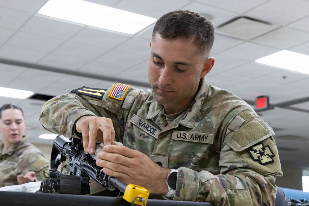 A U.S. Army soldier named Varcoe is performing weapon maintenance or assembly/disassembly on what appears to be an M4/M16-style rifle in an indoor facility. The soldier is wearing OCP (Operational Camouflage Pattern) uniform with a U.S. flag patch and what appears to be a 4th Infantry Division patch on the right sleeve. A second female soldier in the background appears to be engaged in a similar or related task.