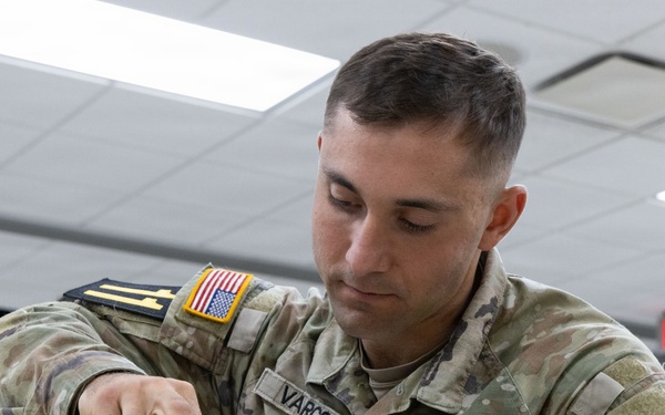 A U.S. Army soldier named Varcoe is performing weapon maintenance or assembly/disassembly on what appears to be an M4/M16-style rifle in an indoor facility. The soldier is wearing OCP (Operational Camouflage Pattern) uniform with a U.S. flag patch and what appears to be a 4th Infantry Division patch on the right sleeve. A second female soldier in the background appears to be engaged in a similar or related task.