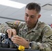 A U.S. Army soldier named Varcoe is performing weapon maintenance or assembly/disassembly on what appears to be an M4/M16-style rifle in an indoor facility. The soldier is wearing OCP (Operational Camouflage Pattern) uniform with a U.S. flag patch and what appears to be a 4th Infantry Division patch on the right sleeve. A second female soldier in the background appears to be engaged in a similar or related task.