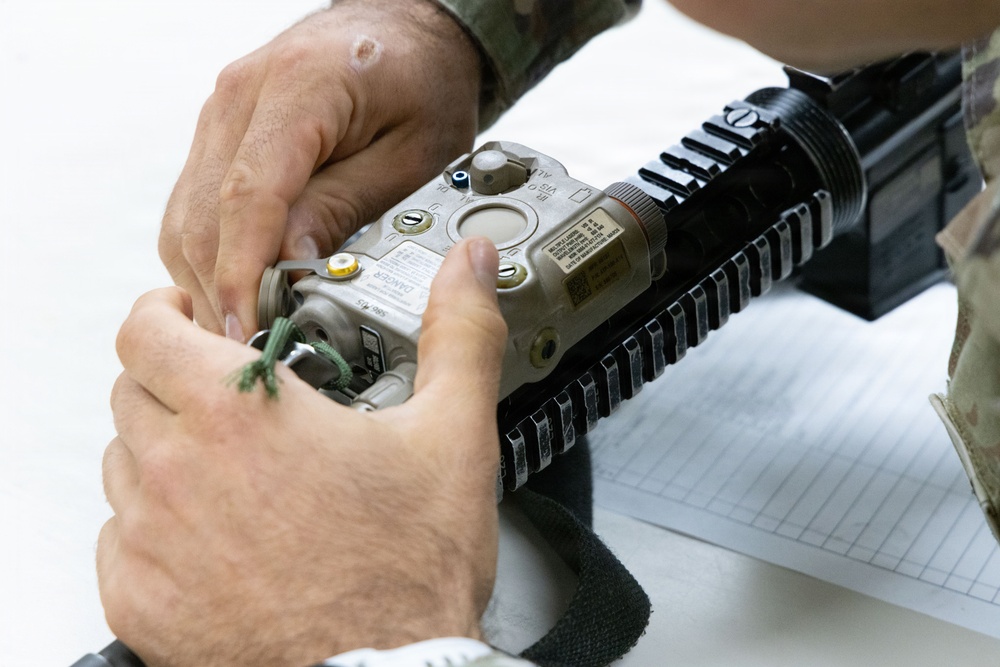 A person in military camouflage uniform is handling a laser aiming device (appears to be an AN/PEQ-15 or similar ATPIAL unit) mounted on a rifle's Picatinny rail system. The individual appears to be performing maintenance, installation, or inspection of the device. A notepad is visible in the background, suggesting a training or equipment check context.