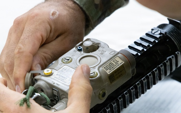 A person in military camouflage uniform is handling a laser aiming device (appears to be an AN/PEQ-15 or similar ATPIAL unit) mounted on a rifle's Picatinny rail system. The individual appears to be performing maintenance, installation, or inspection of the device. A notepad is visible in the background, suggesting a training or equipment check context.