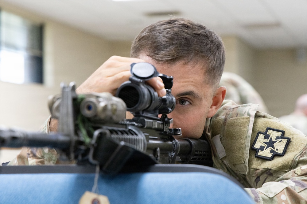 A soldier in U.S. Army Operational Camouflage Pattern (OCP) uniform is aiming down the scope of a rifle in what appears to be an indoor training environment. The soldier wears a unit patch on the right shoulder consistent with the 2nd Infantry Division. The setting suggests a marksmanship training or qualification exercise conducted indoors.
