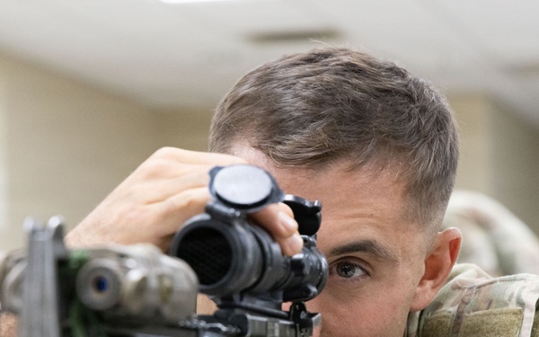 A soldier in U.S. Army Operational Camouflage Pattern (OCP) uniform is aiming down the scope of a rifle in what appears to be an indoor training environment. The soldier wears a unit patch on the right shoulder consistent with the 2nd Infantry Division. The setting suggests a marksmanship training or qualification exercise conducted indoors.