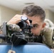 A soldier in U.S. Army Operational Camouflage Pattern (OCP) uniform is aiming down the scope of a rifle in what appears to be an indoor training environment. The soldier wears a unit patch on the right shoulder consistent with the 2nd Infantry Division. The setting suggests a marksmanship training or qualification exercise conducted indoors.
