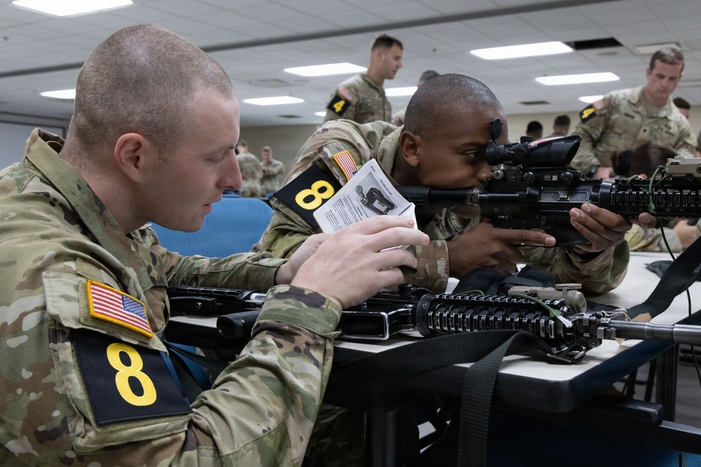 Two U.S. Army soldiers wearing MultiCam (OCP) uniforms and numbered placards are engaged in a weapons training exercise in a classroom setting. The soldier on the left is reading from an instructional document while the soldier on the right is looking through the optic sight of an M4/M16-style rifle. Additional soldiers are visible in the background performing similar activities.