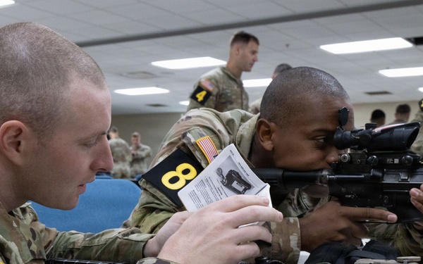 Two U.S. Army soldiers wearing MultiCam (OCP) uniforms and numbered placards are engaged in a weapons training exercise in a classroom setting. The soldier on the left is reading from an instructional document while the soldier on the right is looking through the optic sight of an M4/M16-style rifle. Additional soldiers are visible in the background performing similar activities.