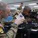 Two U.S. Army soldiers wearing MultiCam (OCP) uniforms and numbered placards are engaged in a weapons training exercise in a classroom setting. The soldier on the left is reading from an instructional document while the soldier on the right is looking through the optic sight of an M4/M16-style rifle. Additional soldiers are visible in the background performing similar activities.