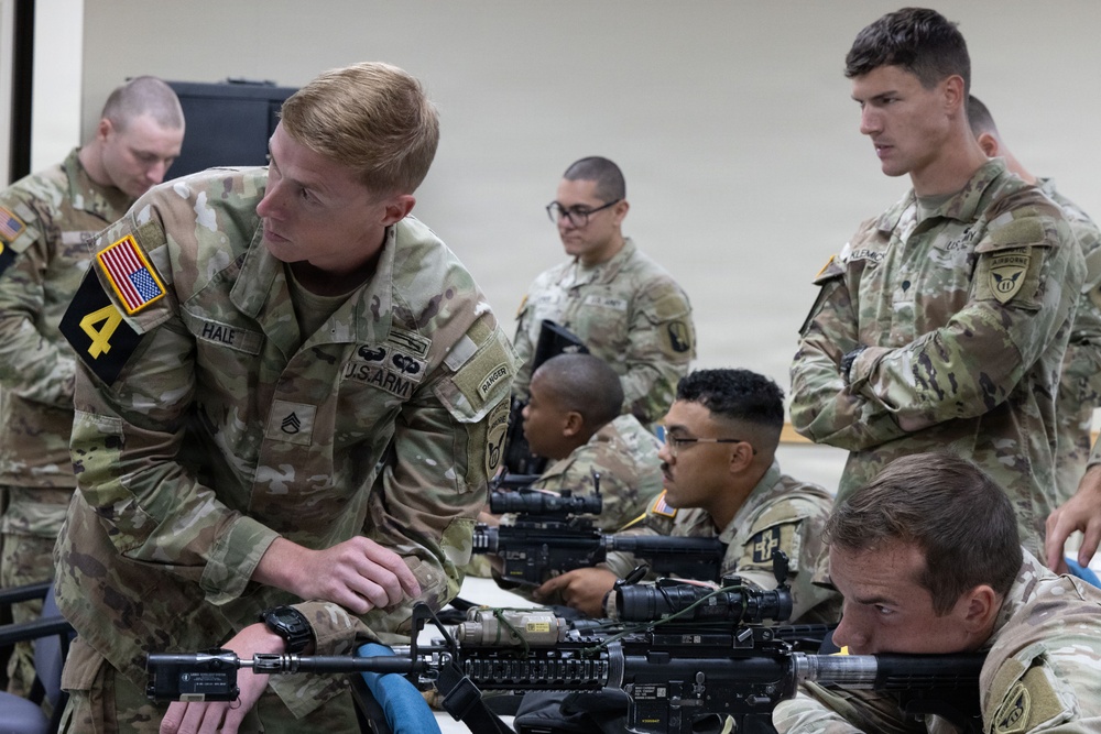 U.S. Army soldiers are engaged in an indoor weapons training or qualification session. A staff sergeant identified as 'HALE' from the 4th Infantry Division is leaning over a table instructing soldiers on rifle handling. Multiple soldiers with M4/M16-style rifles are present in what appears to be a classroom or training bay environment.