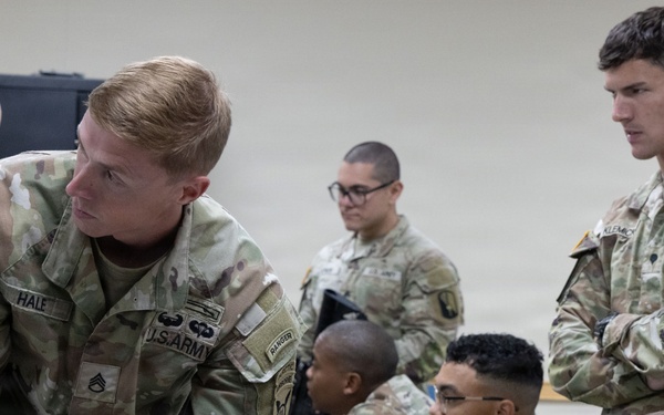 U.S. Army soldiers are engaged in an indoor weapons training or qualification session. A staff sergeant identified as 'HALE' from the 4th Infantry Division is leaning over a table instructing soldiers on rifle handling. Multiple soldiers with M4/M16-style rifles are present in what appears to be a classroom or training bay environment.