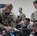 U.S. Army soldiers are engaged in an indoor weapons training or qualification session. A staff sergeant identified as 'HALE' from the 4th Infantry Division is leaning over a table instructing soldiers on rifle handling. Multiple soldiers with M4/M16-style rifles are present in what appears to be a classroom or training bay environment.