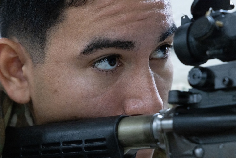 A close-up photograph of a young male individual aiming down the scope of a rifle or long-barreled firearm. The image is tightly framed on his face and the weapon's optic sight, showing intense focus. The subject appears to be in a training or shooting context.
