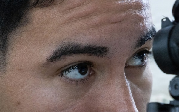 A close-up photograph of a young male individual aiming down the scope of a rifle or long-barreled firearm. The image is tightly framed on his face and the weapon's optic sight, showing intense focus. The subject appears to be in a training or shooting context.