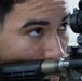 A close-up photograph of a young male individual aiming down the scope of a rifle or long-barreled firearm. The image is tightly framed on his face and the weapon's optic sight, showing intense focus. The subject appears to be in a training or shooting context.