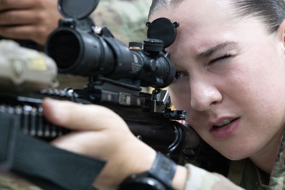 A female soldier in military camouflage uniform is aiming through the optical scope of a rifle. She has her dominant eye pressed against the scope eyepiece while her non-dominant eye is closed, indicating active target acquisition. The close-up shot captures fine details of both the weapon system and the shooter's focused expression.