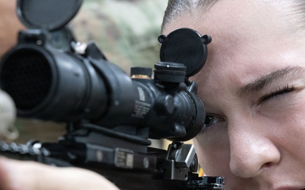 A female soldier in military camouflage uniform is aiming through the optical scope of a rifle. She has her dominant eye pressed against the scope eyepiece while her non-dominant eye is closed, indicating active target acquisition. The close-up shot captures fine details of both the weapon system and the shooter's focused expression.