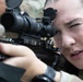 A female soldier in military camouflage uniform is aiming through the optical scope of a rifle. She has her dominant eye pressed against the scope eyepiece while her non-dominant eye is closed, indicating active target acquisition. The close-up shot captures fine details of both the weapon system and the shooter's focused expression.
