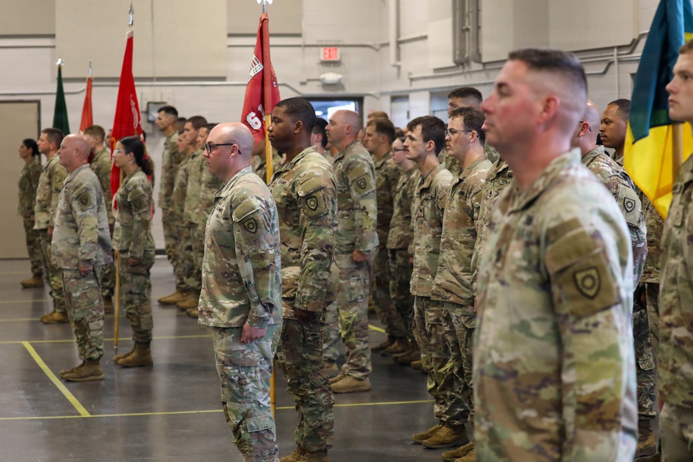 A formation of U.S. Army soldiers in OCP (Operational Camouflage Pattern) uniforms standing at attention inside a large indoor facility, likely a gymnasium or armory. Multiple unit guidons and flags are visible, suggesting a formal military ceremony such as a change of command, deployment ceremony, or unit activation. Soldiers are arranged in organized rows facing a common direction.