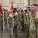 A formation of U.S. Army soldiers in OCP (Operational Camouflage Pattern) uniforms standing at attention inside a large indoor facility, likely a gymnasium or armory. Multiple unit guidons and flags are visible, suggesting a formal military ceremony such as a change of command, deployment ceremony, or unit activation. Soldiers are arranged in organized rows facing a common direction.