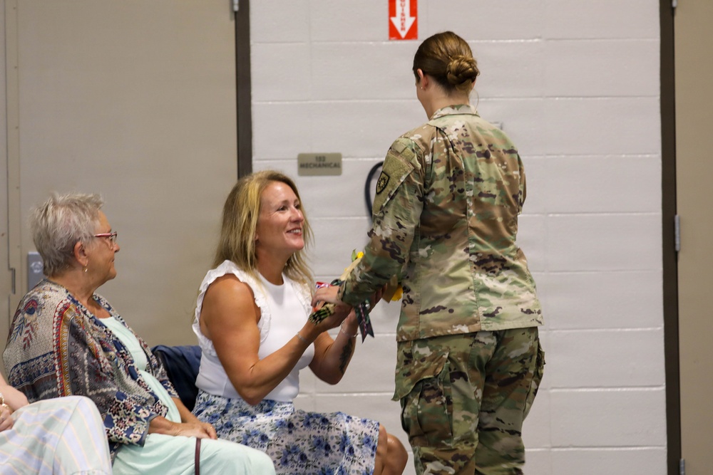 A female soldier in Army OCP (Operational Camouflage Pattern) uniform is presenting or pinning something to a smiling civilian woman seated in a chair. An older woman seated nearby observes the interaction with a smile. The setting appears to be an indoor military ceremony or event, likely a deployment, homecoming, or recognition ceremony.